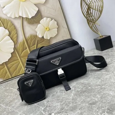 A black nylon camera bag with a small matching pouch clipped to its strap, standing on gray marble in front of a carved white flower panel.