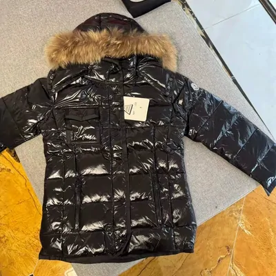A glossy black puffer with a brown fur-trimmed hood, laid open on a gray mat over a yellow floor, its paper tag still attached.
